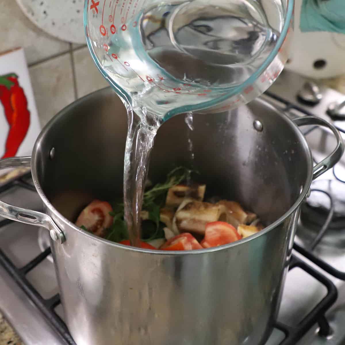 A person pouring water into a large stock pot that is filled with roasted beef marrow bones, herbs, and sautéed vegetables.