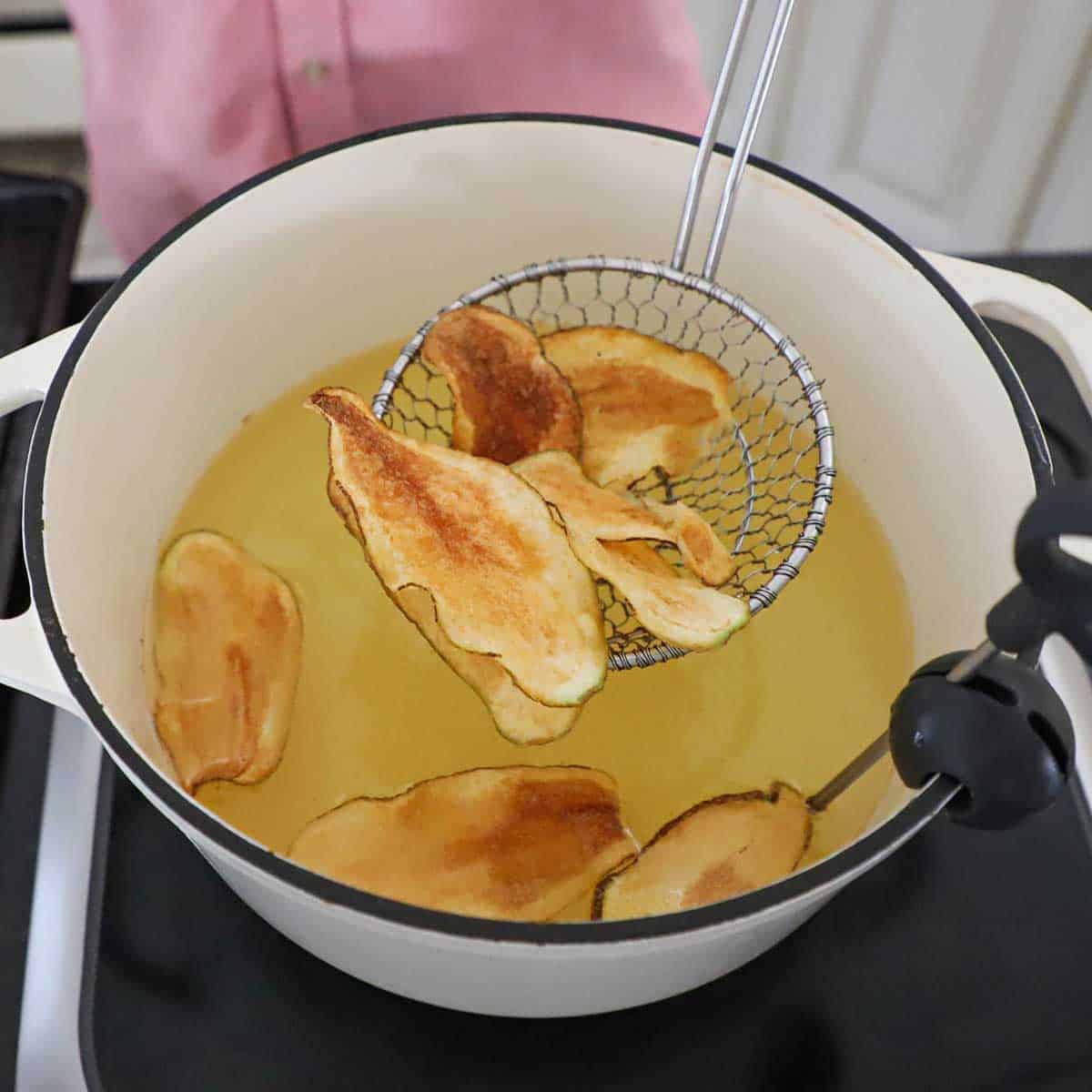A person using a metal spider to lift crispy homemade potato chips out of a white pot that is filled with hot oil.
