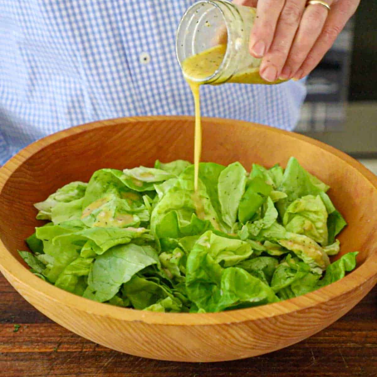 A person pouring a homemade French vinaigrette over torn butter lettuce in a large wooden salad bowl.