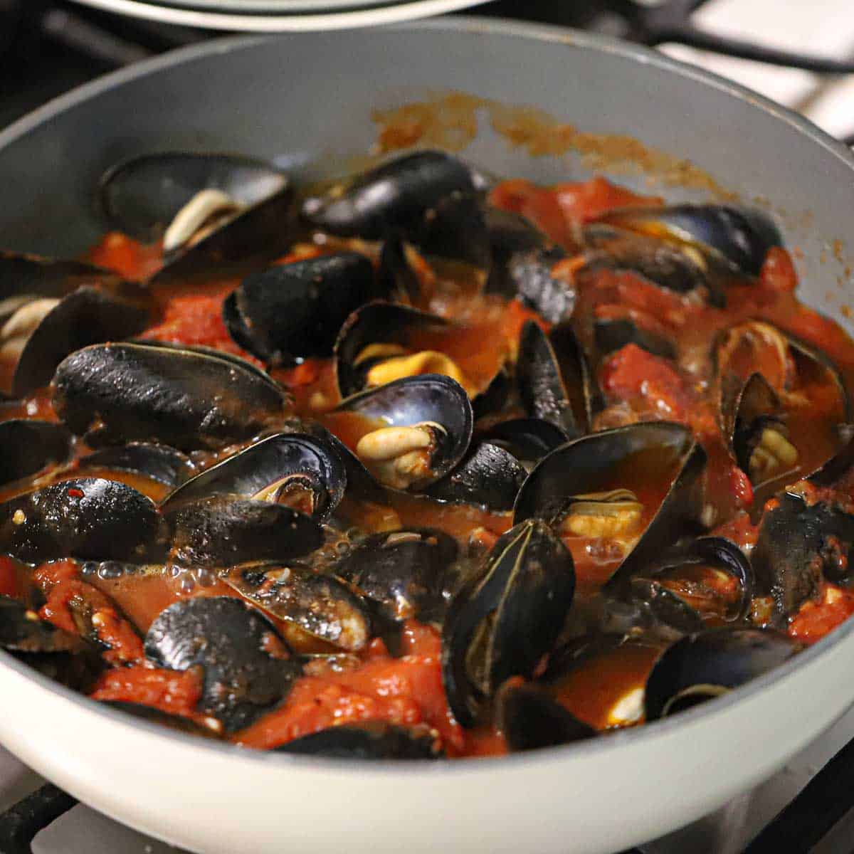 A close-up view of mussels that are open and revealing the meat inside all simmering in a marinara sauce in a large skillet.