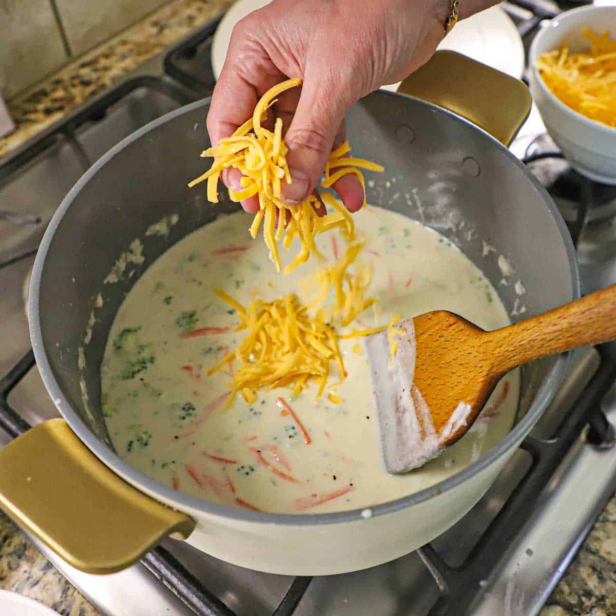A person using his hand to sprinkle shredded cheddar cheese into a pot filled with broccoli cheddar soup.