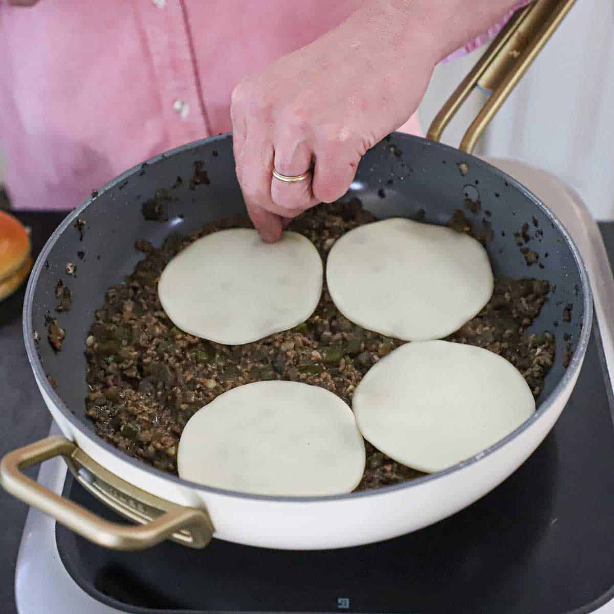 A person placing circular slices of Provolone cheese into over the surface of simmering Philly cheesesteak sloppy Joes in a large skillet.