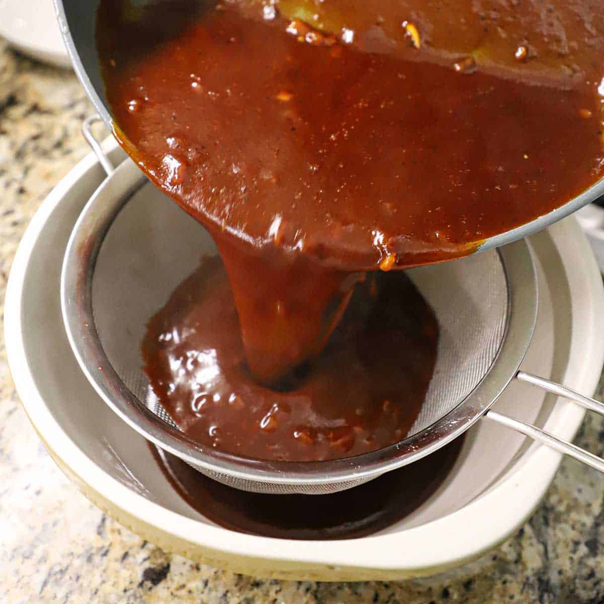 A person pouring homemade easy BBQ sauce from a large skillet into a fine-mesh sieve over a large ceramic bowl.