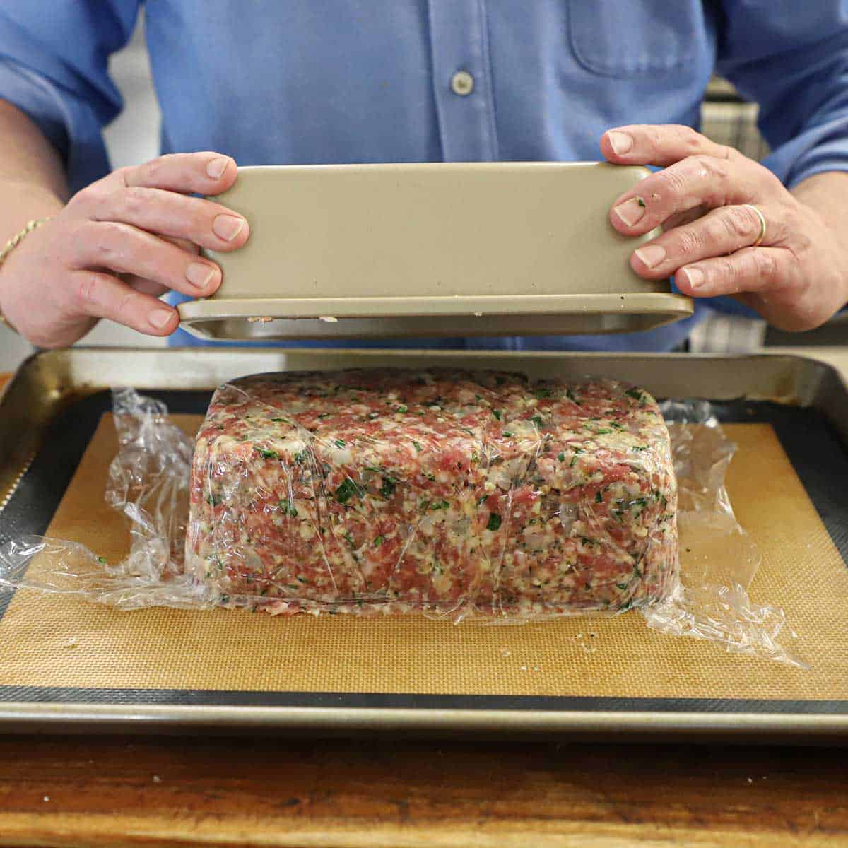 A person lifting a metal loaf pan from a loaf of uncooked Italian meatloaf on a baking sheet lined with a silicone matt.