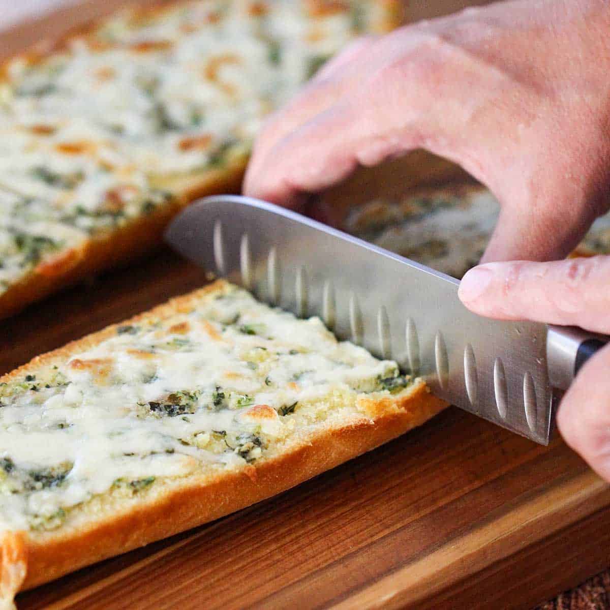 A person using a large chef's knife to cut a freshly baked loaf of cheesy garlic bread in half on a wooden cutting board.