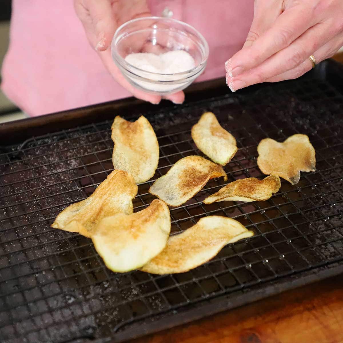 A person using his fingers to sprinkle fine sea salt over the surface of freshly fried crispy homemade potato chips that are resting on a baking rack inside a baking sheet.