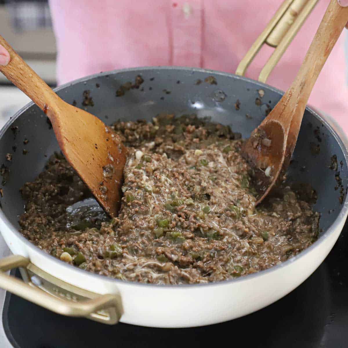A person using two large wooden utensils to stir melted Provolone cheese into a skillet filled with Philly cheesesteak sloppy Joes.