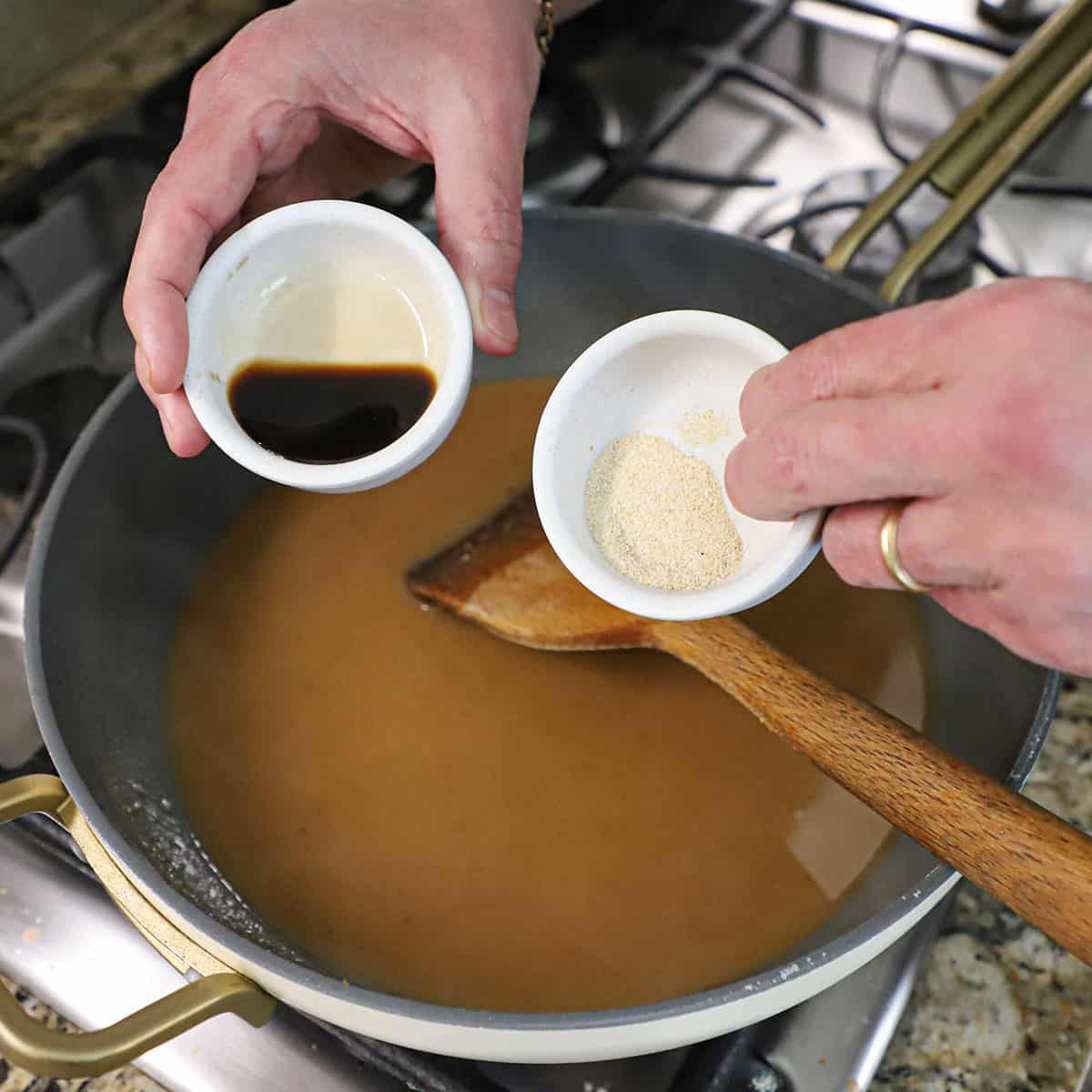 A person holding a small white bowl filled with 1 teaspoon of garlic powder in one hand and holding another small white bowl holding 1 teaspoon of Worcestershire sauce both over a skillet filled with a brown gravy.