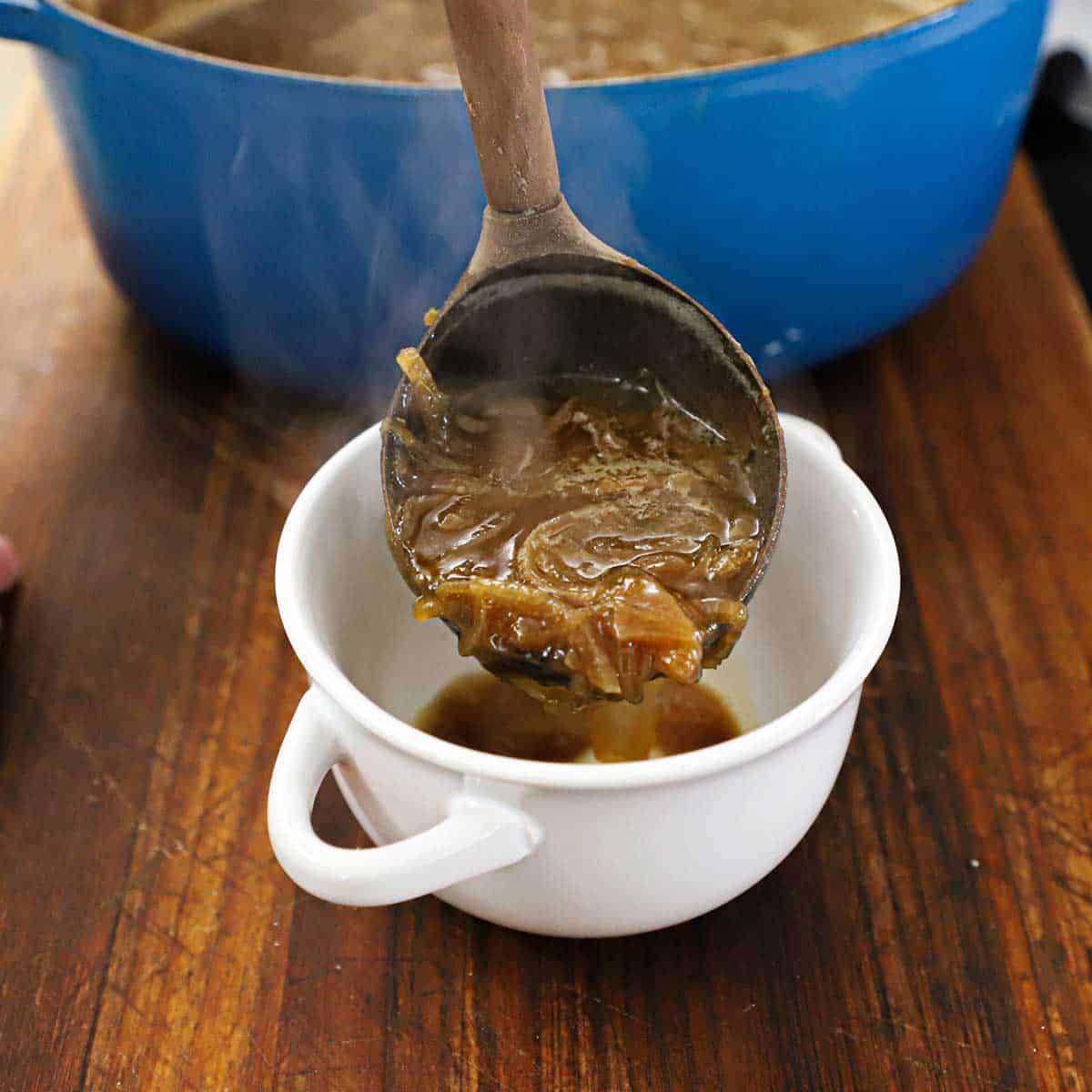 A person using a large wooden ladle to transfer homemade French onion soup from a large Dutch oven into a white soup bowl.