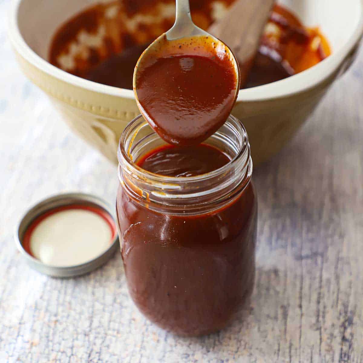A person transferring homemade easy BBQ sauce from a large spoon into a glass jar that is filled with the sauce.