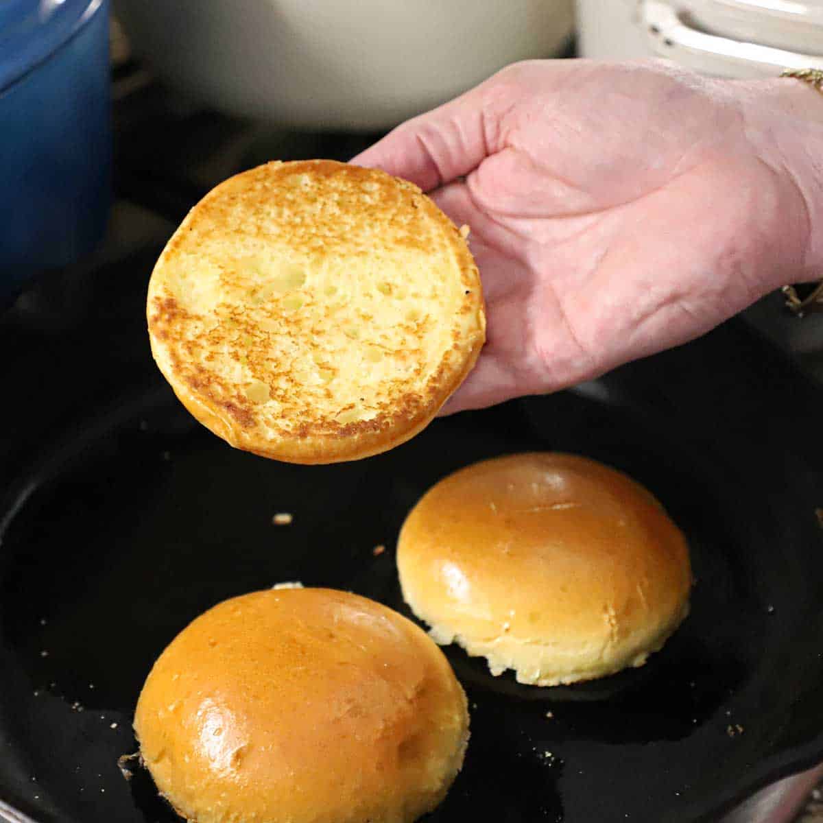A person holding a lightly toasted brioche bun that was toasted, along with other buns, on a hot cast-iron skillet.