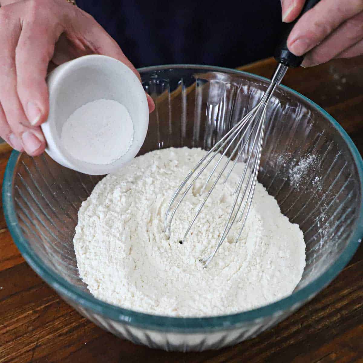 A person using a whisk to combine flour in a glass bowl with baking powder and salt.