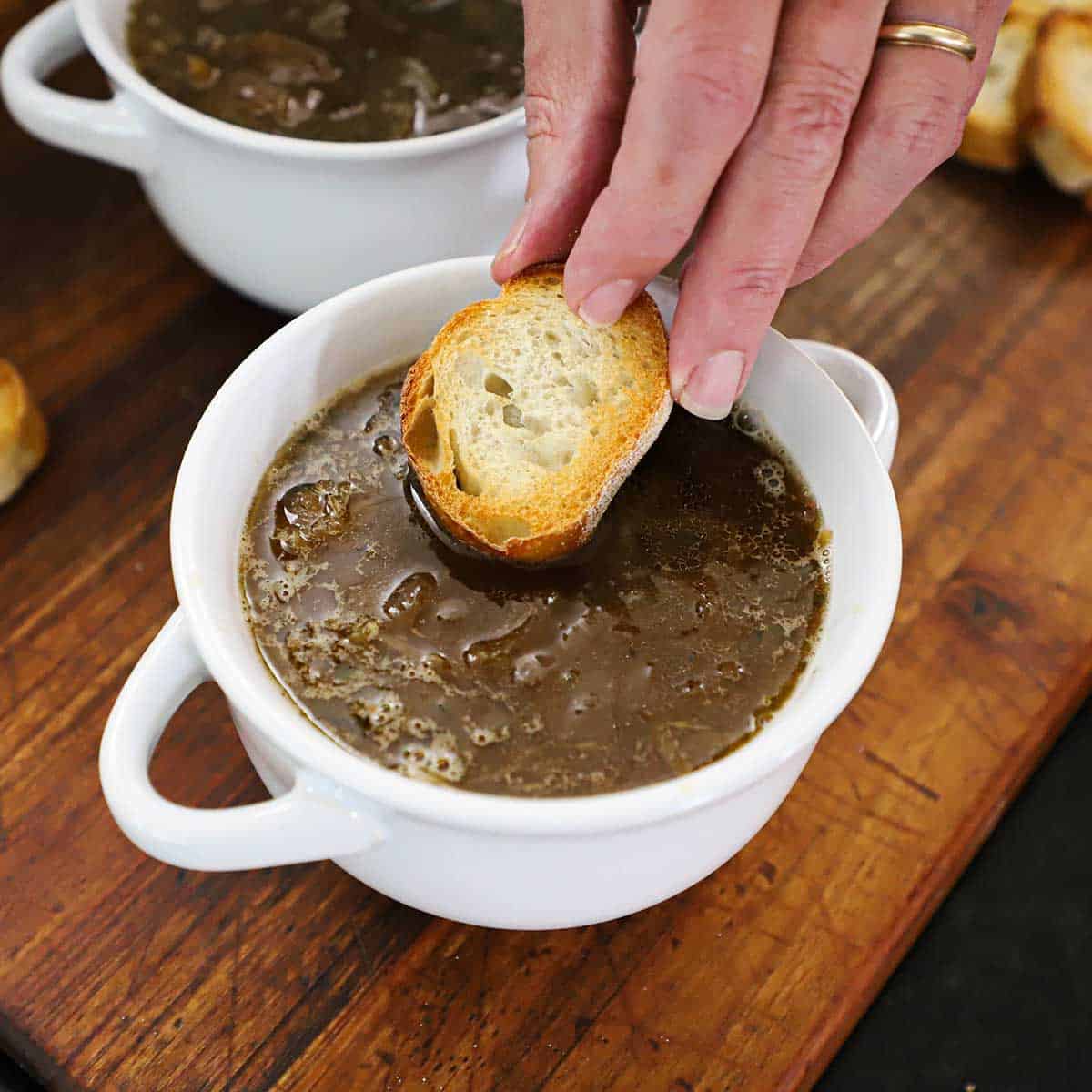 A person placing a slice of toasted baguette over the top of homemade French onion soup in a white soup bowl.