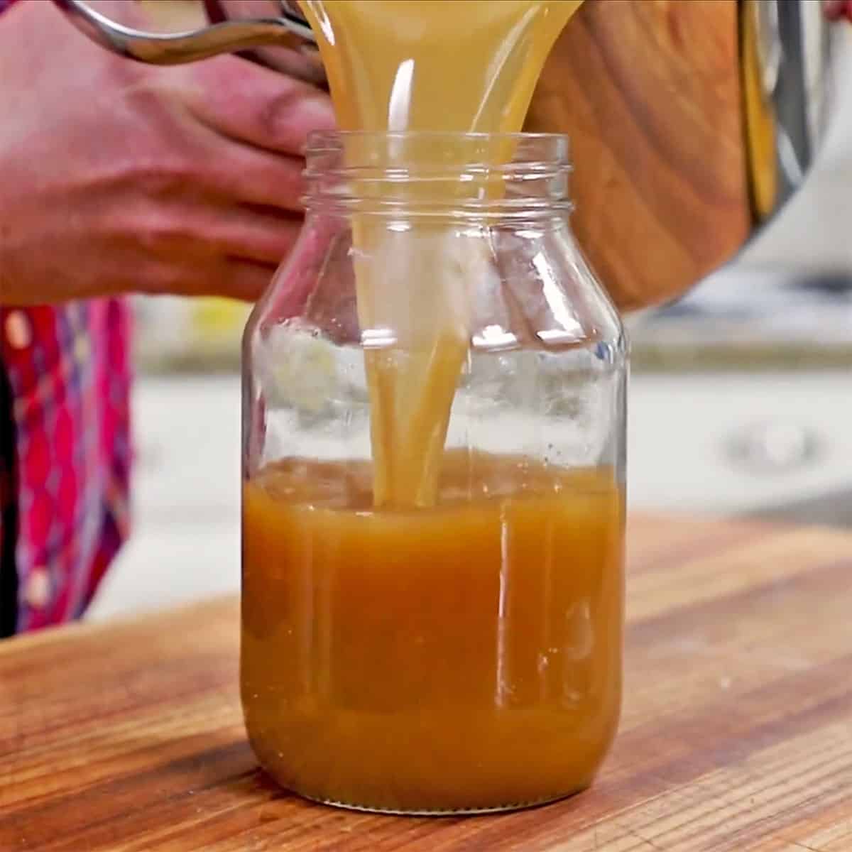 A person pouring homemade beef stock from a stock pot into a large glass jar resting on a wooden cutting board.