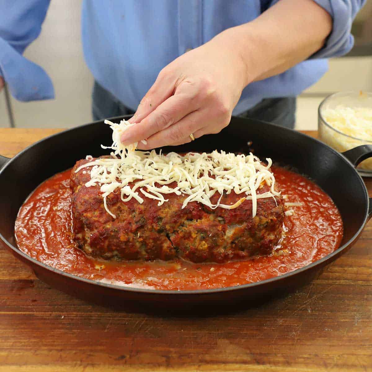 A person sprinkling shredded mozzarella onto a partially cooked Italian-style meatloaf that is resting in a pan partially filled with marinara sauce.