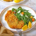 A close-up view of a dinner plate filled with a serving of crispy chicken Milanese alongside an arugula salad with a couple of lemon wedges next to it.