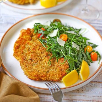 A close-up view of a dinner plate filled with a serving of crispy chicken Milanese alongside an arugula salad with a couple of lemon wedges next to it.