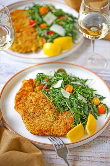 Two dinner plates filled a serving of crispy chicken Milanese with an arugula salad next to the cutlet along with two lemon wedges.