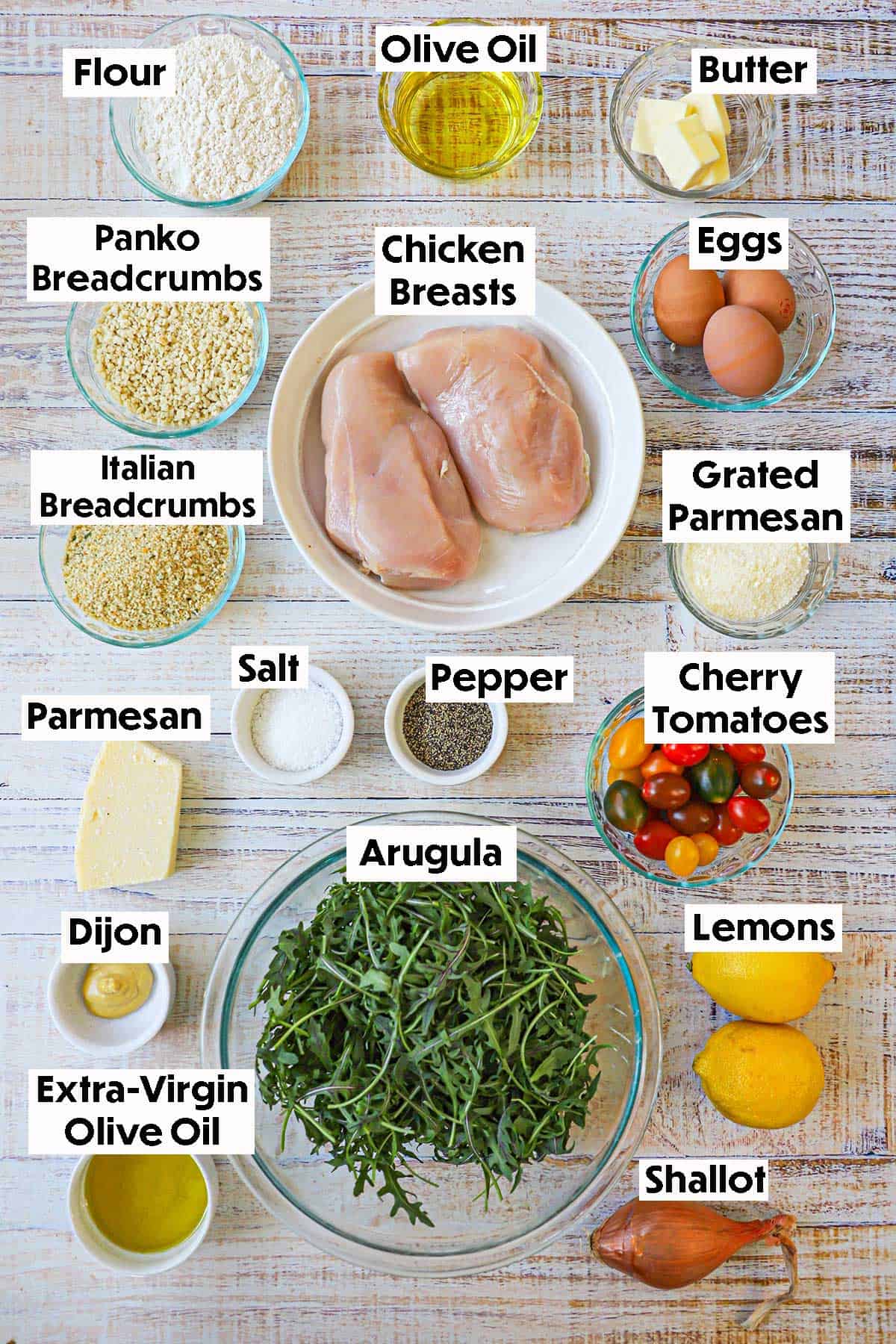 An arrangement of ingredients for crispy chicken Milanese on a grey wooden background including chicken breasts, breadcrumbs, flour, eggs, olive oil, butter, arugula, tomatoes, shallots, lemons, Parmesan cheese, and spices.