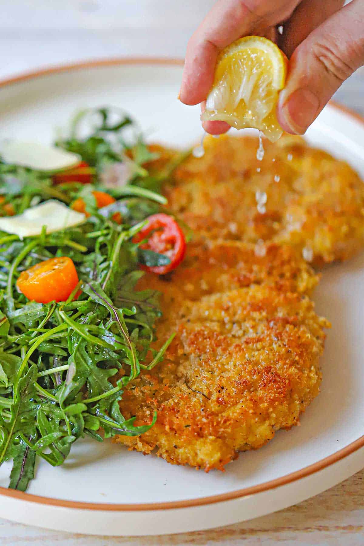 A person squeezing a lemon wedge over a crispy chicken Milanese cutlet next to an arugula salad with cherry tomatoes.