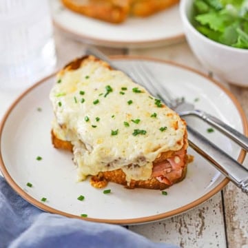 A close-up view of two plates both filled with a freshly prepared Essential Croque Monsieur with a bowl filled with a green salad nearby.