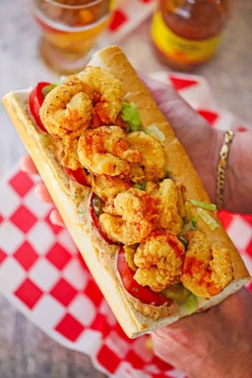 A person using two hands to hold a large New Orleans Fried Shrimp Po-boy over a basket lined with red and white checkered wax paper.