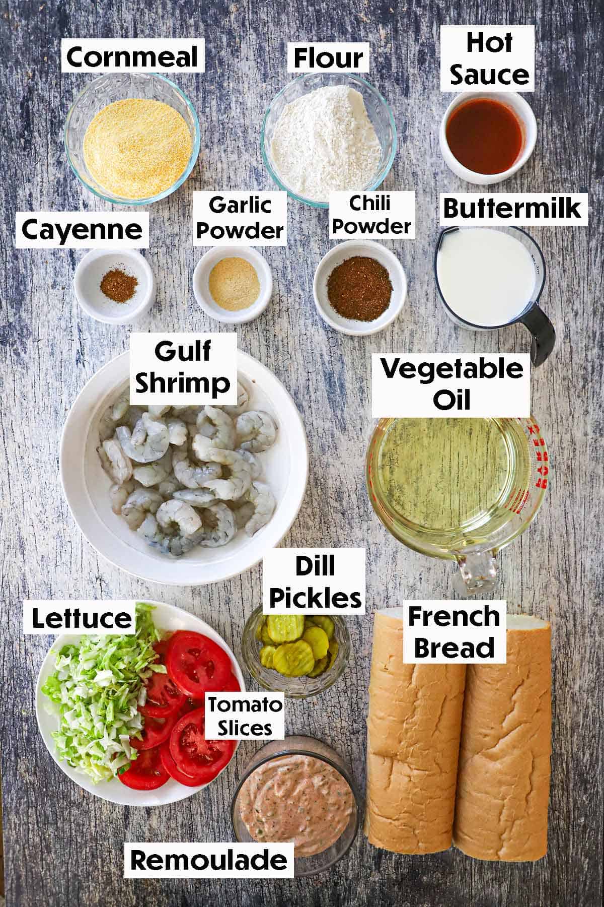 An arrangement of ingredients for New Orleans Fried Shrimp Po-boy on a grey wooden background including raw gulf shrimp, cornmeal, flour, buttermilk, oil, French bread, remoulade, pickles, lettuce, tomatoes, and seasonings.