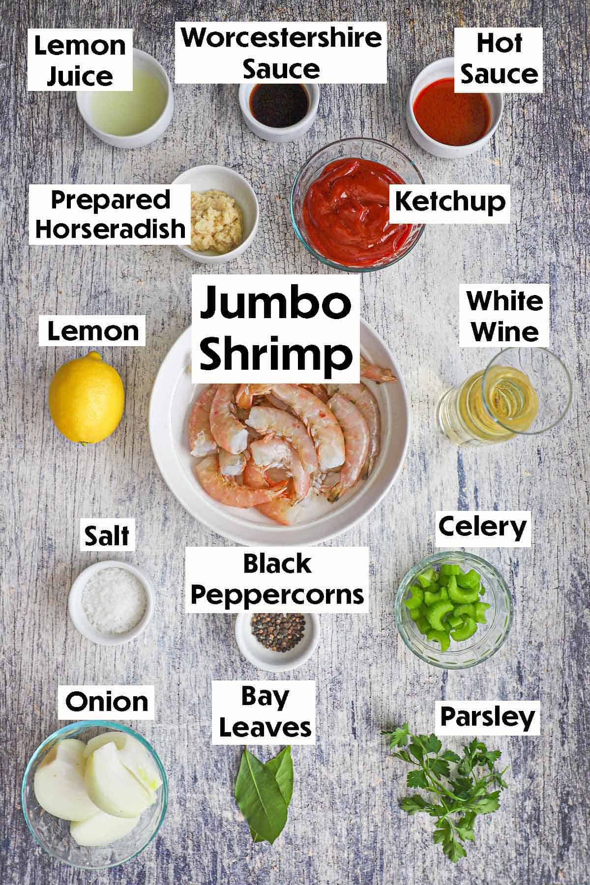 An arrangement of ingredients for Classic Jumbo Shrimp Cocktail on a grey wooden background including uncooked shrimp, lemon juice, Worcestershire sauce, ketchup, horseradish, wine, lemon, celery, onion, herbs, and seasonings.