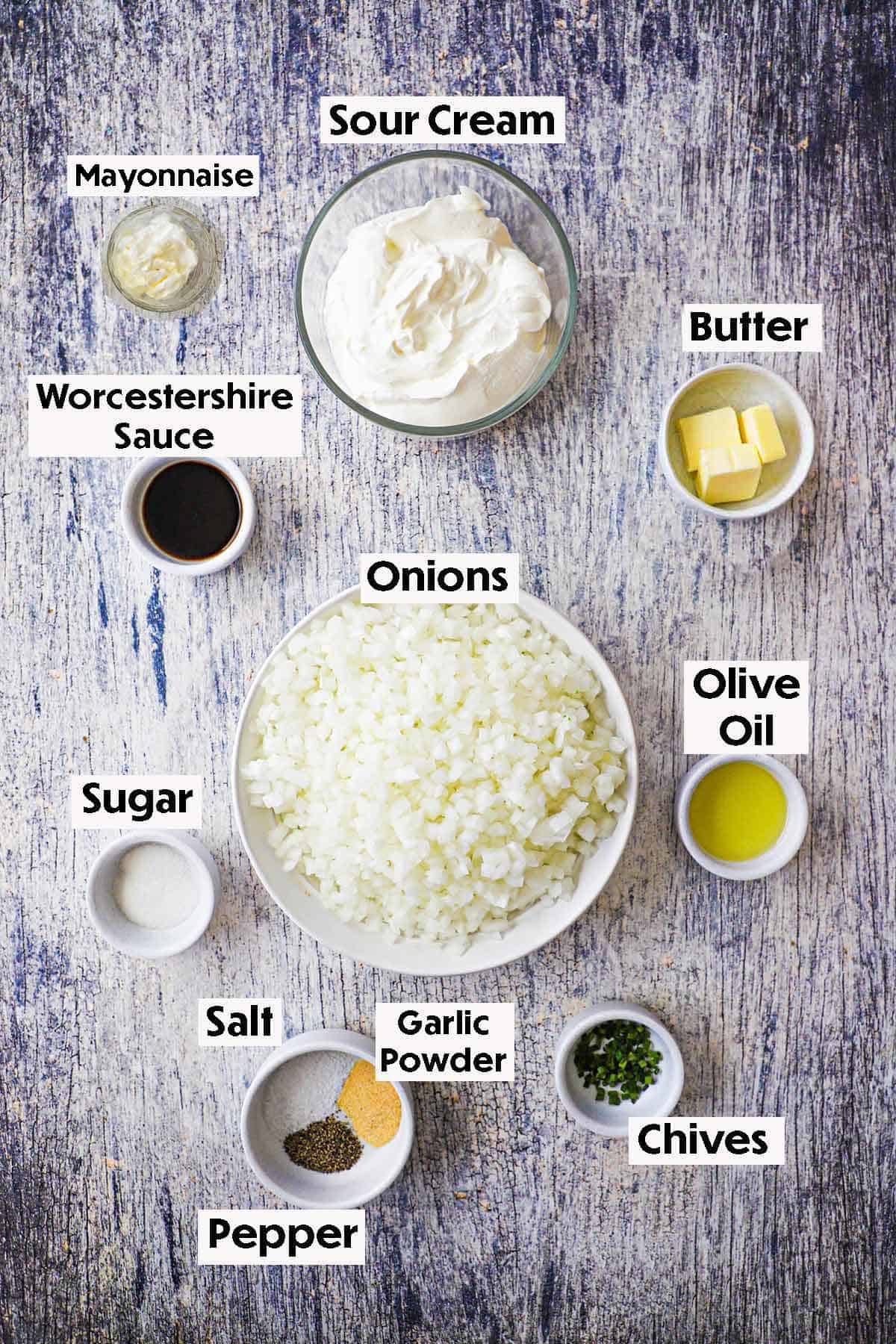 An arrangement of ingredients for homemade French onion dip on a grey wooden background including chopped onions, olive oil, butter, sour cream, mayonnaise, Worcestershire sauce, sugar, and spices.