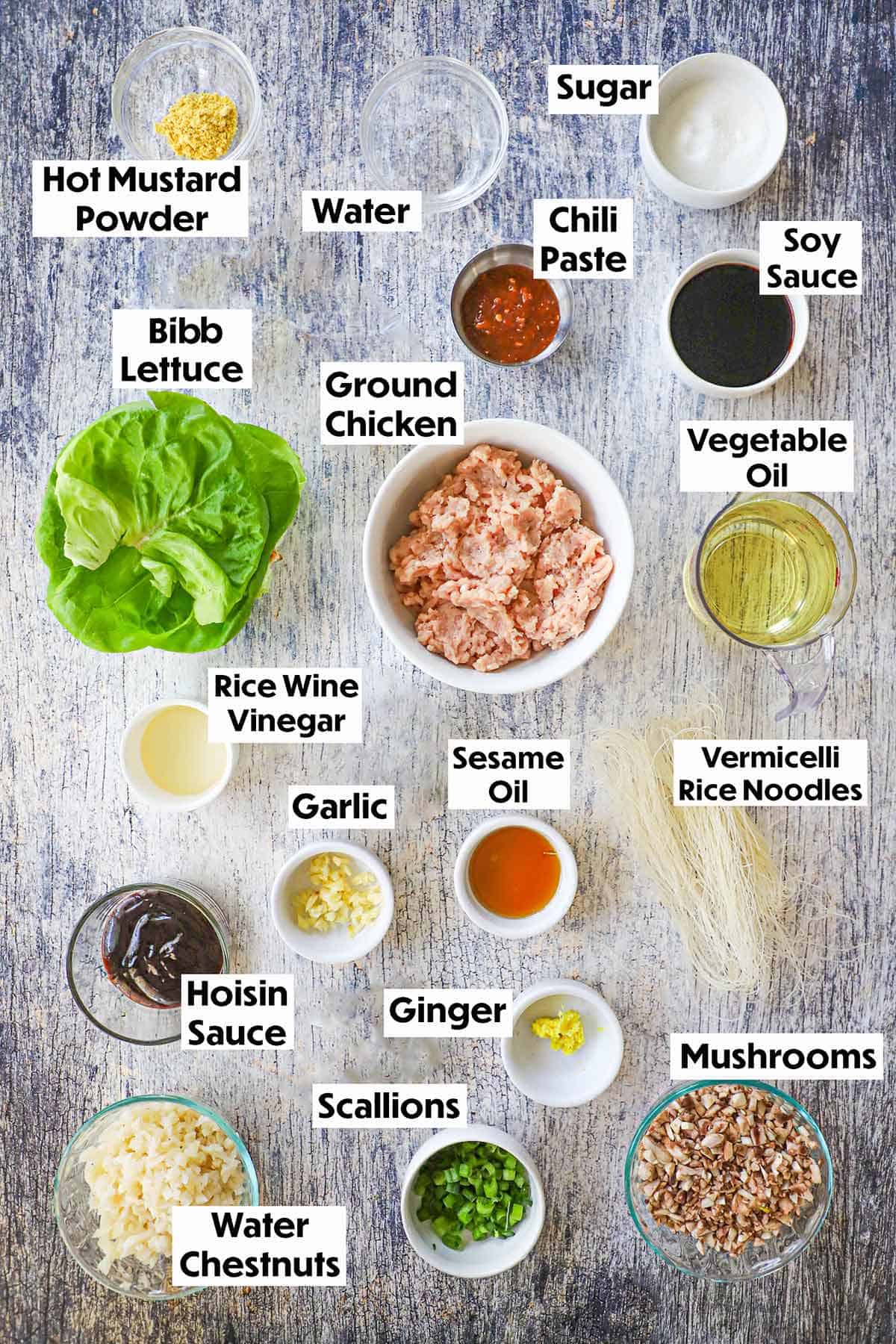 An arrangement of ingredients for P.F. Chang's Chicken Lettuce Wraps Copycat Recipe including groudn chicken, Bibb, lettuce, soy sauce, rice wine vinegar, rice vermicelli, oil, hosing, water chestnuts, mushrooms, scallions, and seasonings.