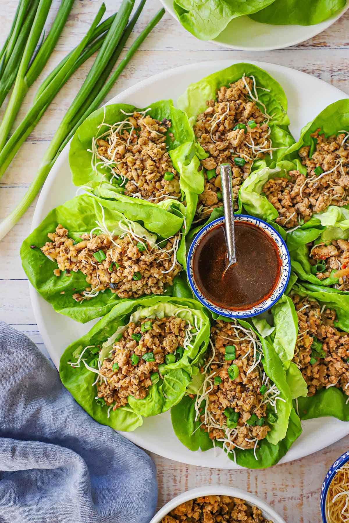 An overhead view of a circular white platter filled with P.F. Chang's Chicken Lettuce Wraps with a festive bowl filled with a zesty red sauce in the middle of the platter.