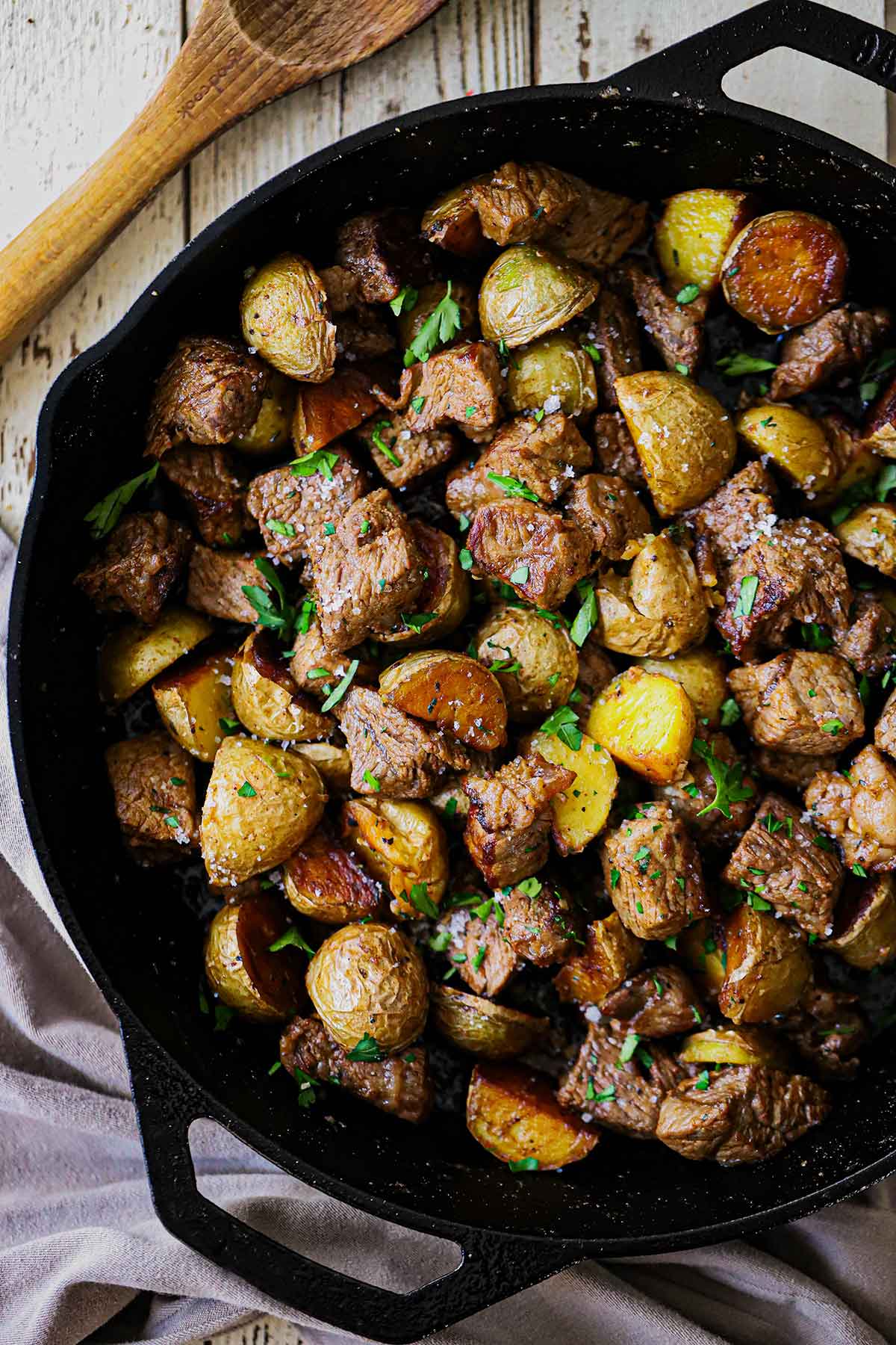 An overhead view of Garlic Butter Steak Bites and Potatoes in a large black cast-iron skillet garnished with chopped Italian parsley and coarse sea salt.
