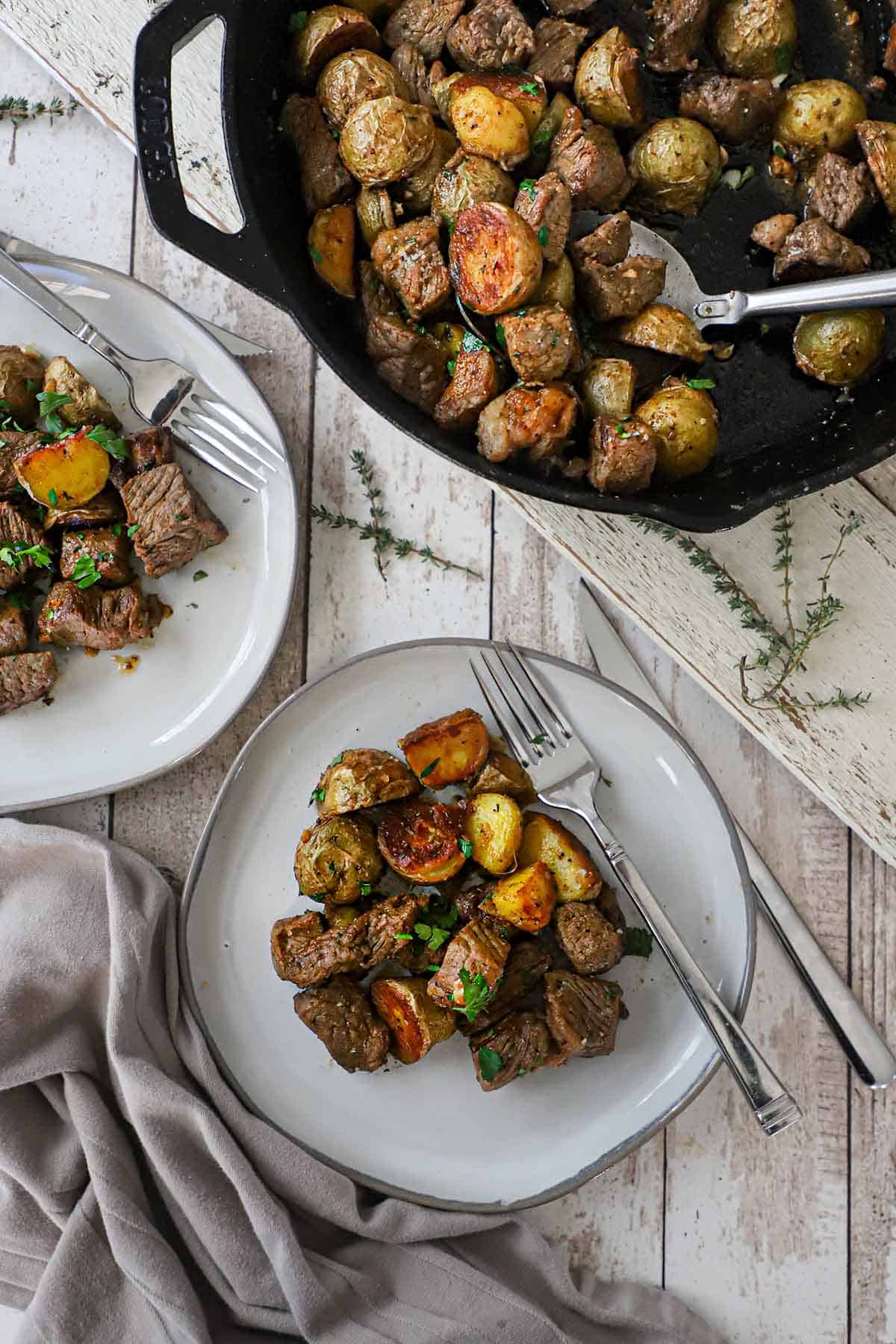 An overhead view of two plates filled with servings of garlic butter steak bites and potatoes with a skillet filled with the same nearby.