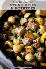 An overhead view of Garlic Butter Steak Bites and Potatoes in a large black cast-iron skillet garnished with chopped Italian parsley and coarse sea salt.