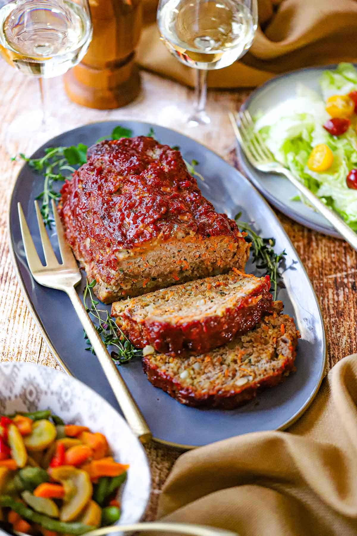 A juicy turkey meatloaf that has two slices cut away resting on an oval blue platter with gold serving fork nearby, as well as two glasses of white wine and a green salad.