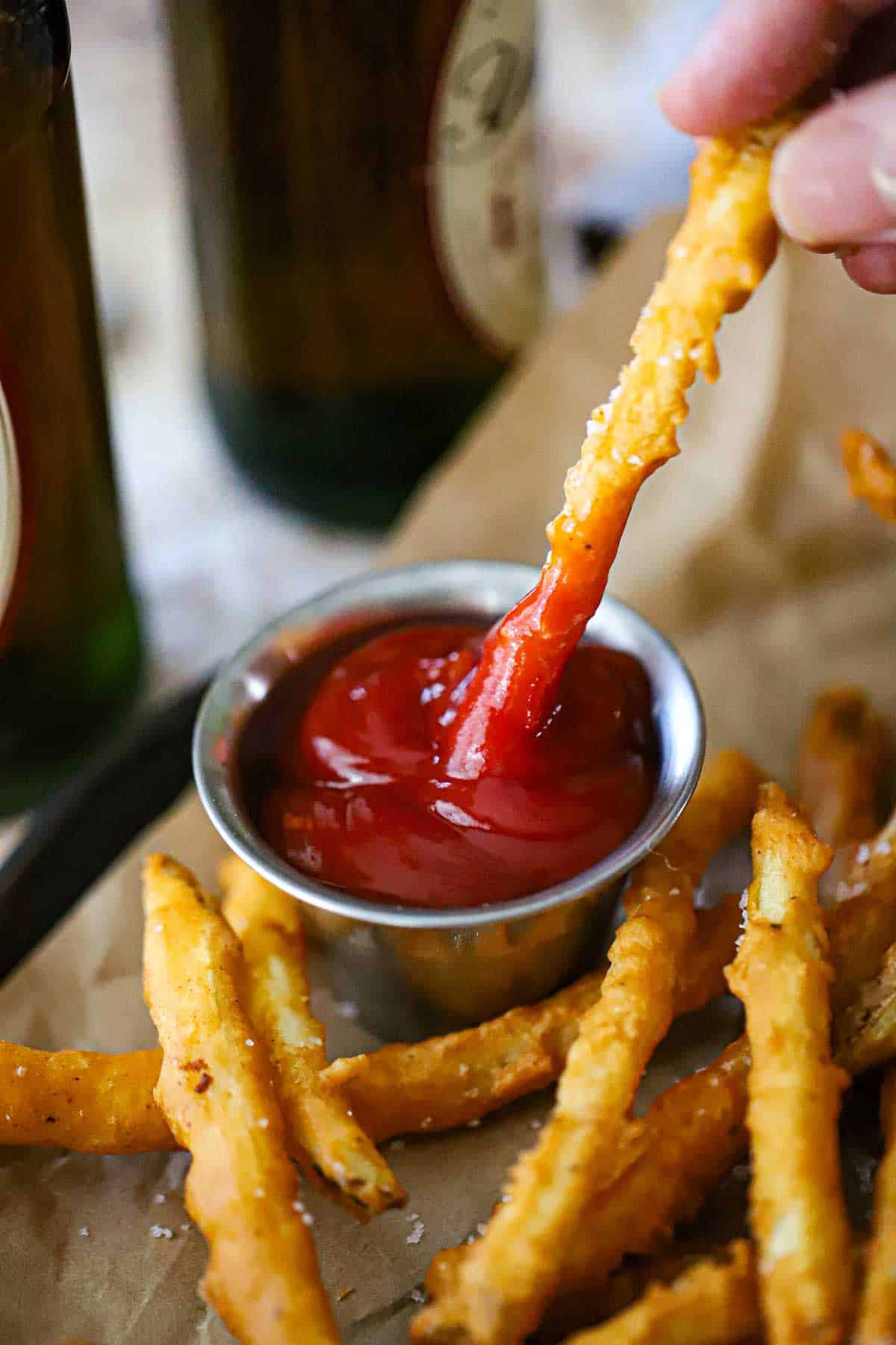 A person plunging the ultimate beer battered French fry into a small condiment vessel filled ketchup next a pile of more French fries.