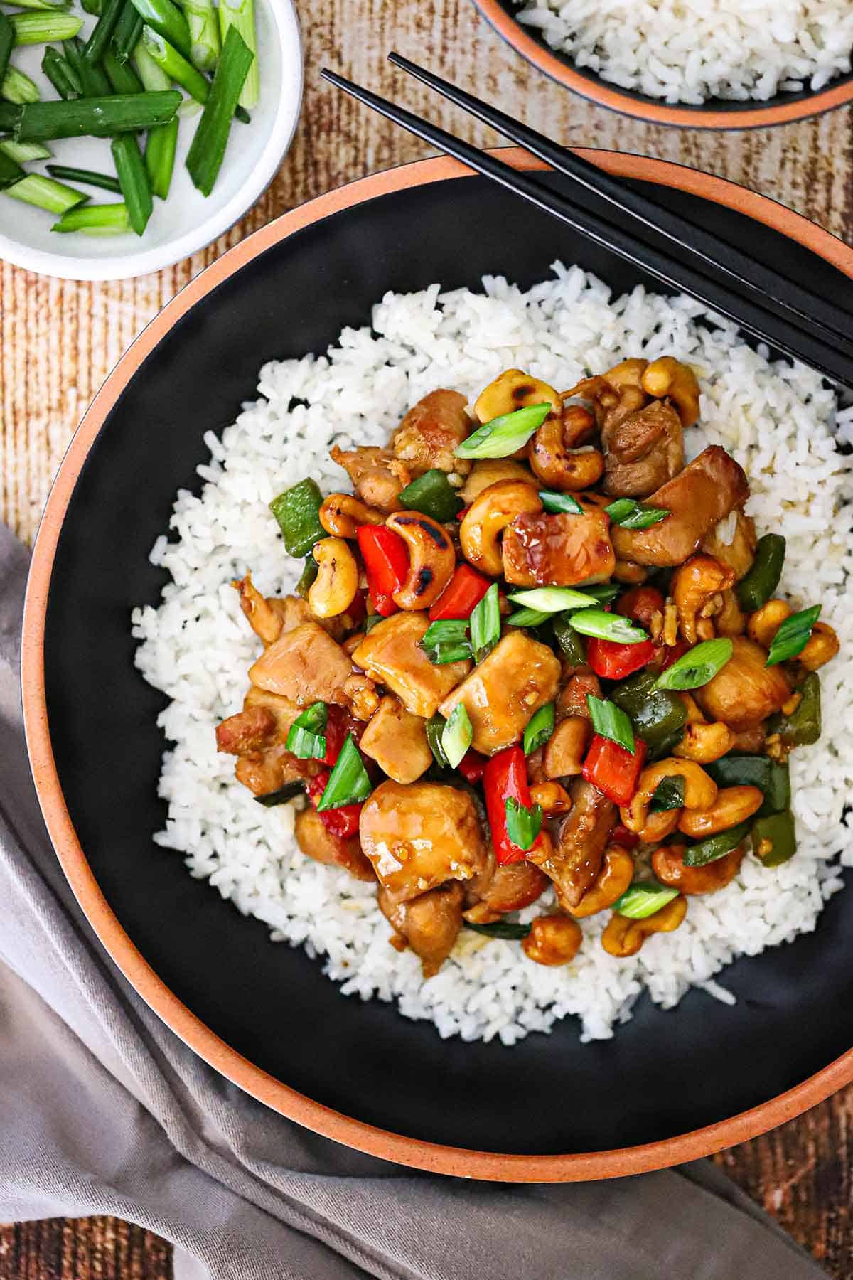 An overhead view of a black bowl filled with a serving of cashew chicken on white rice with two chopsticks resting on the side of the bowl.