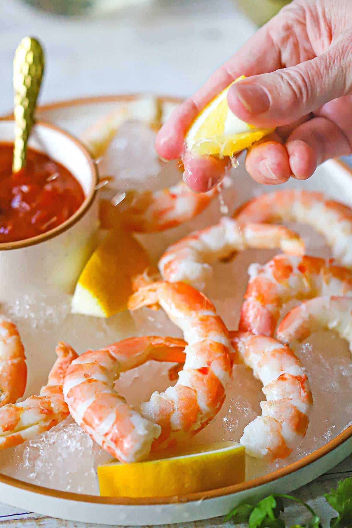 A person squeezing a lemon wedge over a plate filled with Classic Jumbo Shrimp Cocktail.