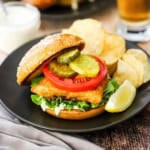A fried fish sandwich on a toasted bun loaded with tartar sauce, lettuce, tomato, and pickles next to potato chips and a lemon wedge all on a black dinner plate.