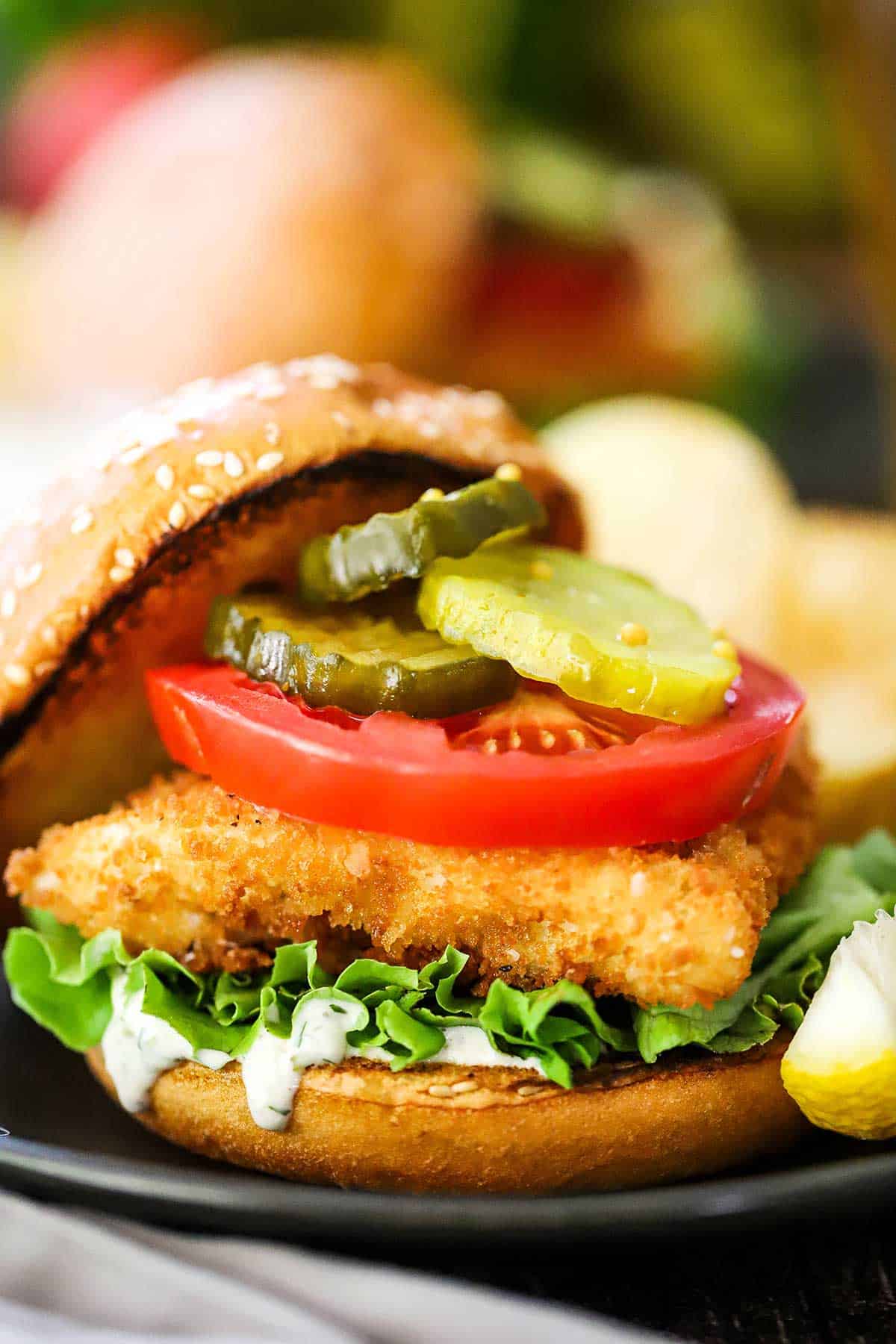 A straight-on view of a fried fish sandwich on a toasted bun with a layer of tartar sauce, green leaf lettuce, and topped with a sliced tomato and pickle chips.