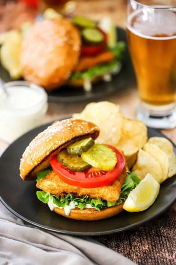 A fried fish sandwich on a toasted bun loaded with tartar sauce, lettuce, tomato, and pickles next to potato chips and a lemon wedge all on a black dinner plate.