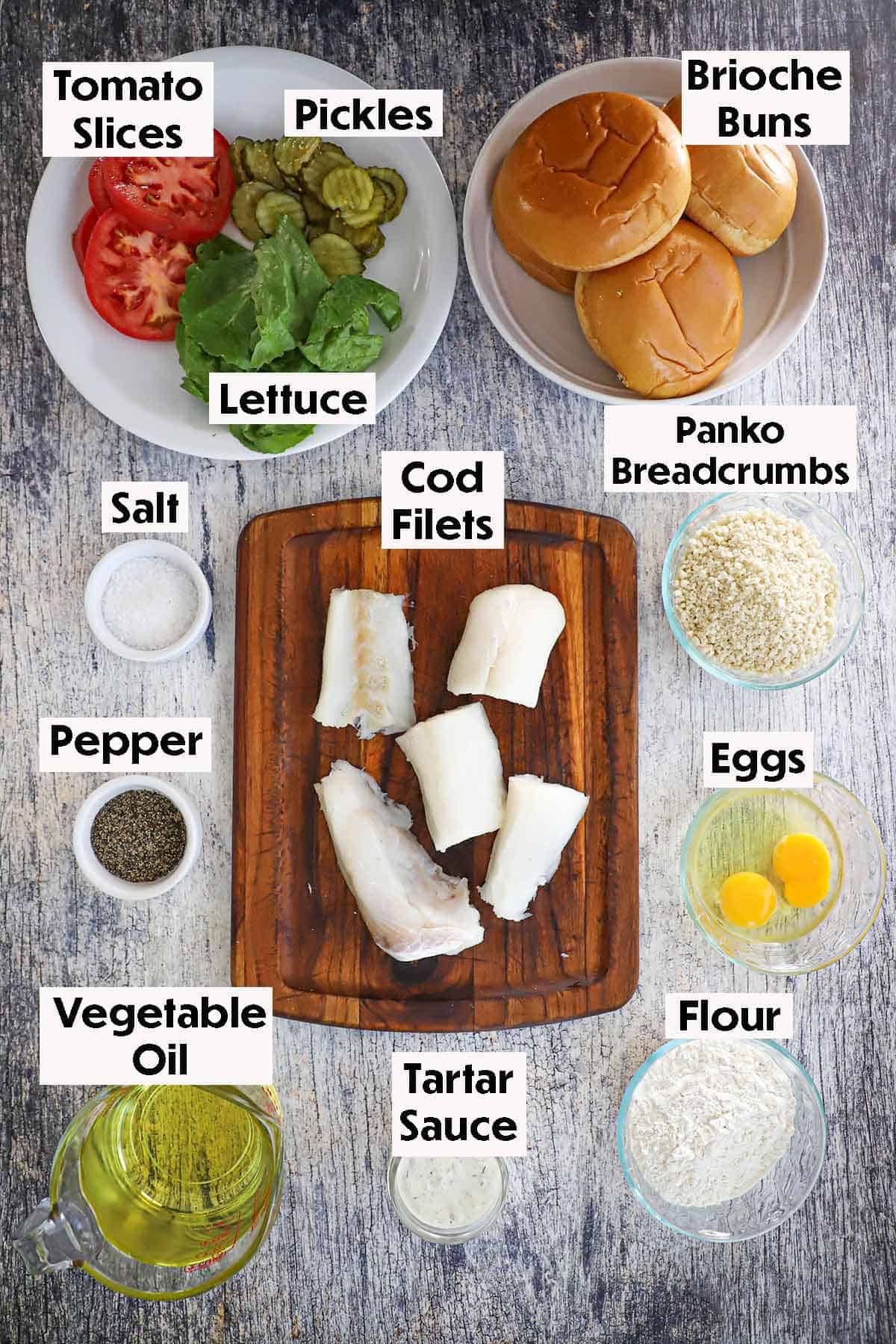 An overhead view of ingredients for fried fish sandwich including cod filets, Panko breadcrumbs, eggs, flour, oil, buns, tomato slices, lettuce, pickle, seasonings, and tartar sauce.