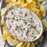 An overhead view of an oval bowl filled with homemade French onion dip garnished with snipped chives on a platter filled with Kettle potato chips.