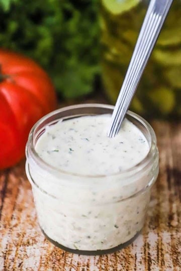 A small glass jar filled to the top with homemade tartar sauce with a spoon in t, and a red tomato, green lettuce, and dill pickles nearby.