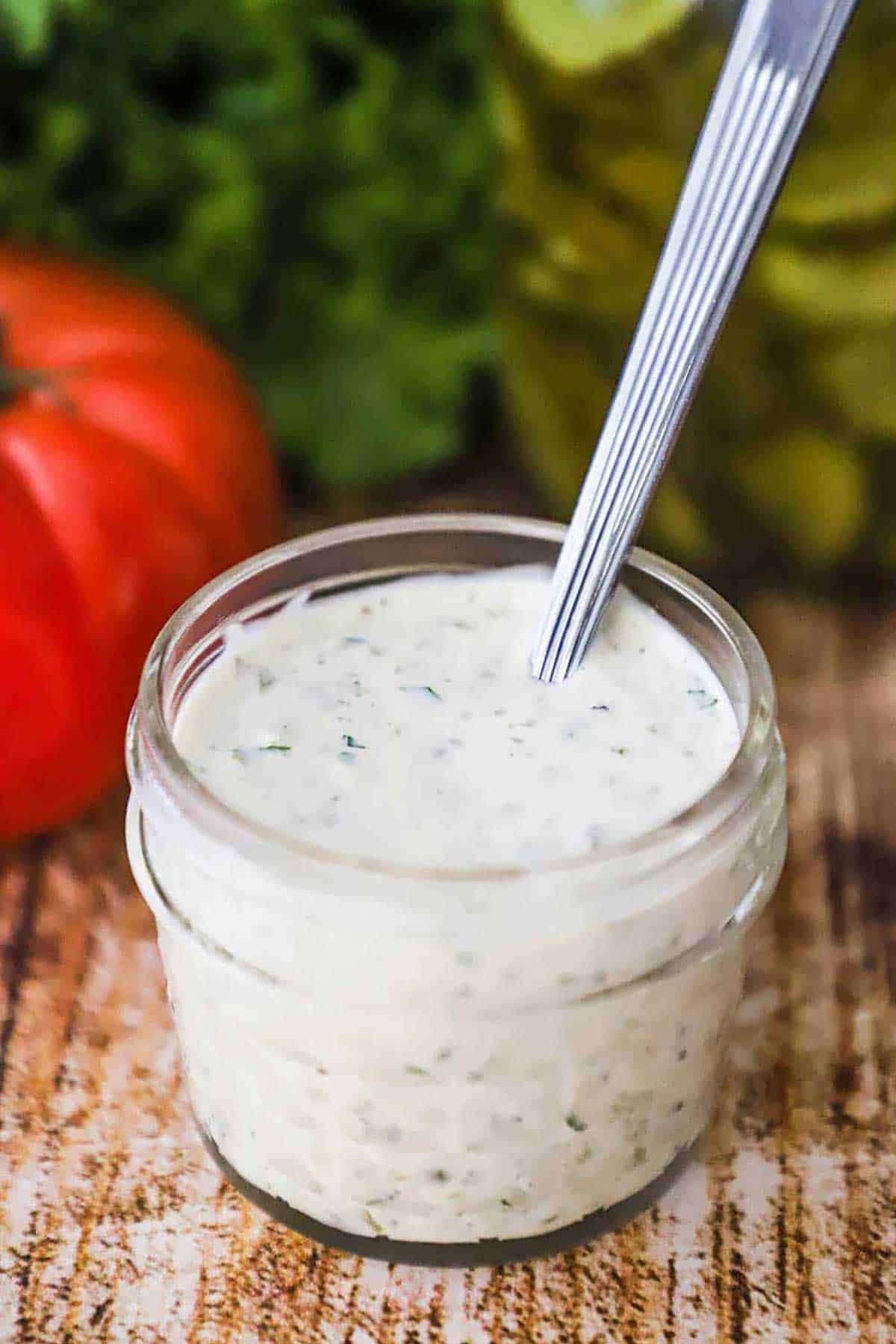 A small glass jar filled to the top with homemade tartar sauce with a spoon in t, and a red tomato, green lettuce, and dill pickles nearby.