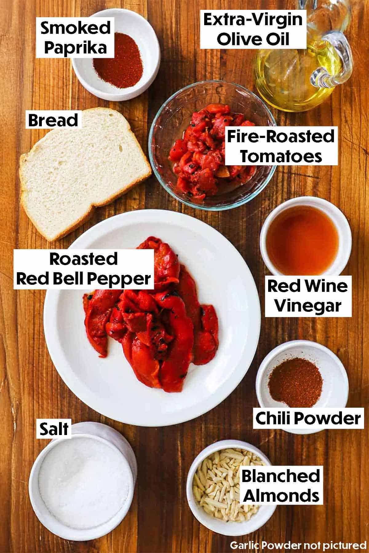 An arrangement of ingredients for easy romesco sauce on a wooden background including roasted red bell peppers, a slice of white bread, diced tomatoes, olive oil, red wine vinegar, almonds, and seasonings.