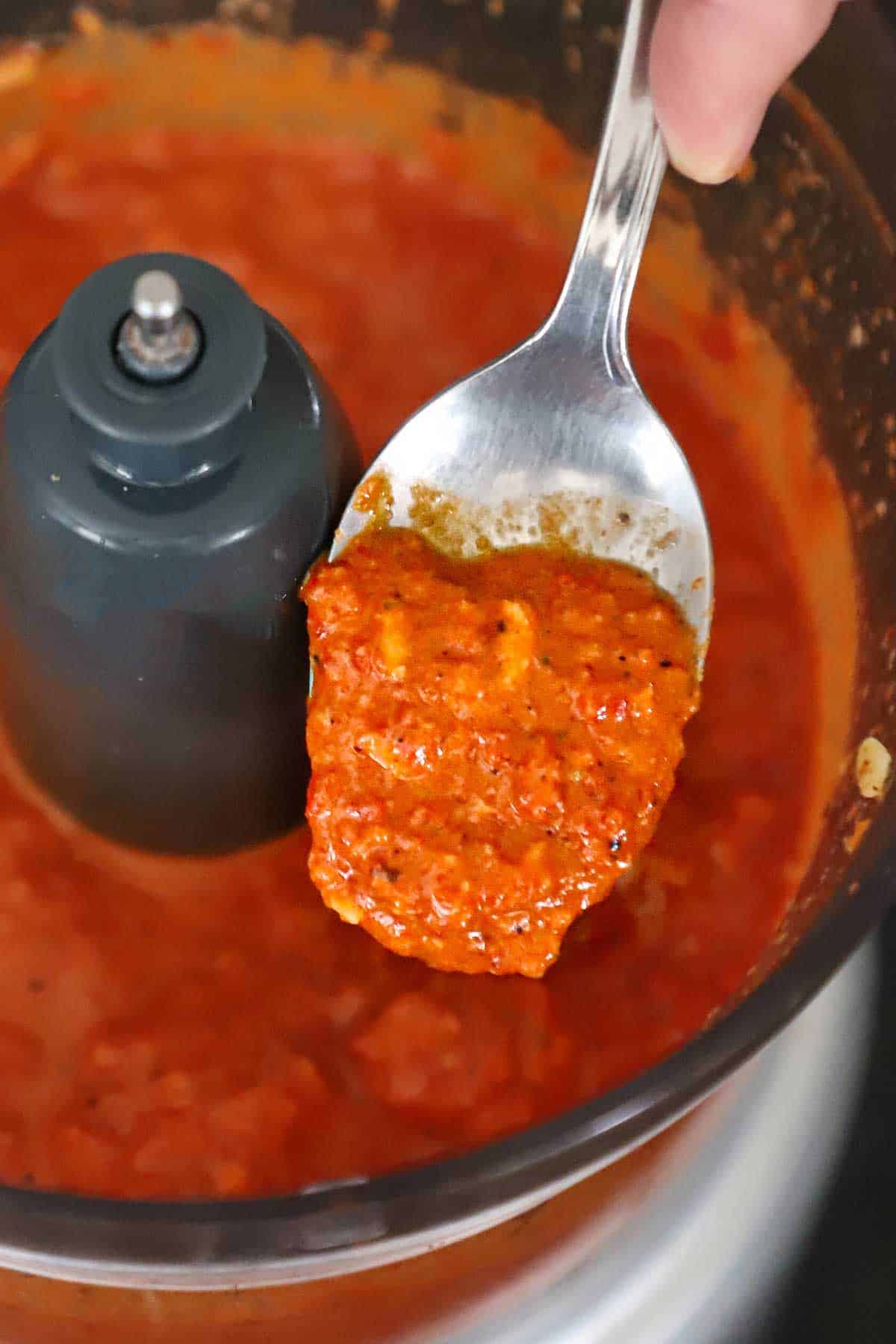 A person using a spoon of easy homemade romesco sauce up from a bowl of a food processor that was just to purée the sauce.