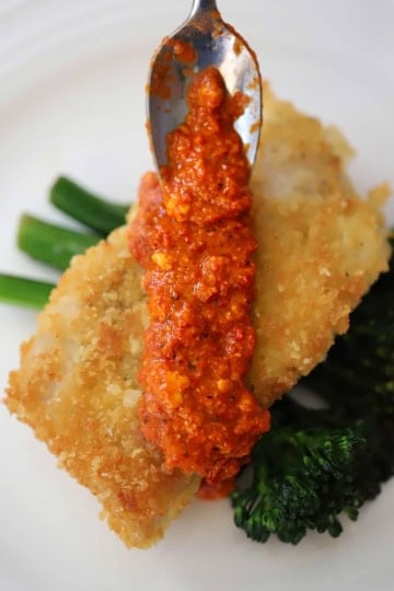 A spoon being used to spread easy homemade romesco sauce of a breaded baked fillet of code resting on two pieces of steamed broccolini.