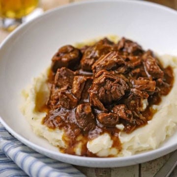 A close-up view of a shallow white bowl filled with slow cooker beef tips and gravy over a pile of mashed potatoes.