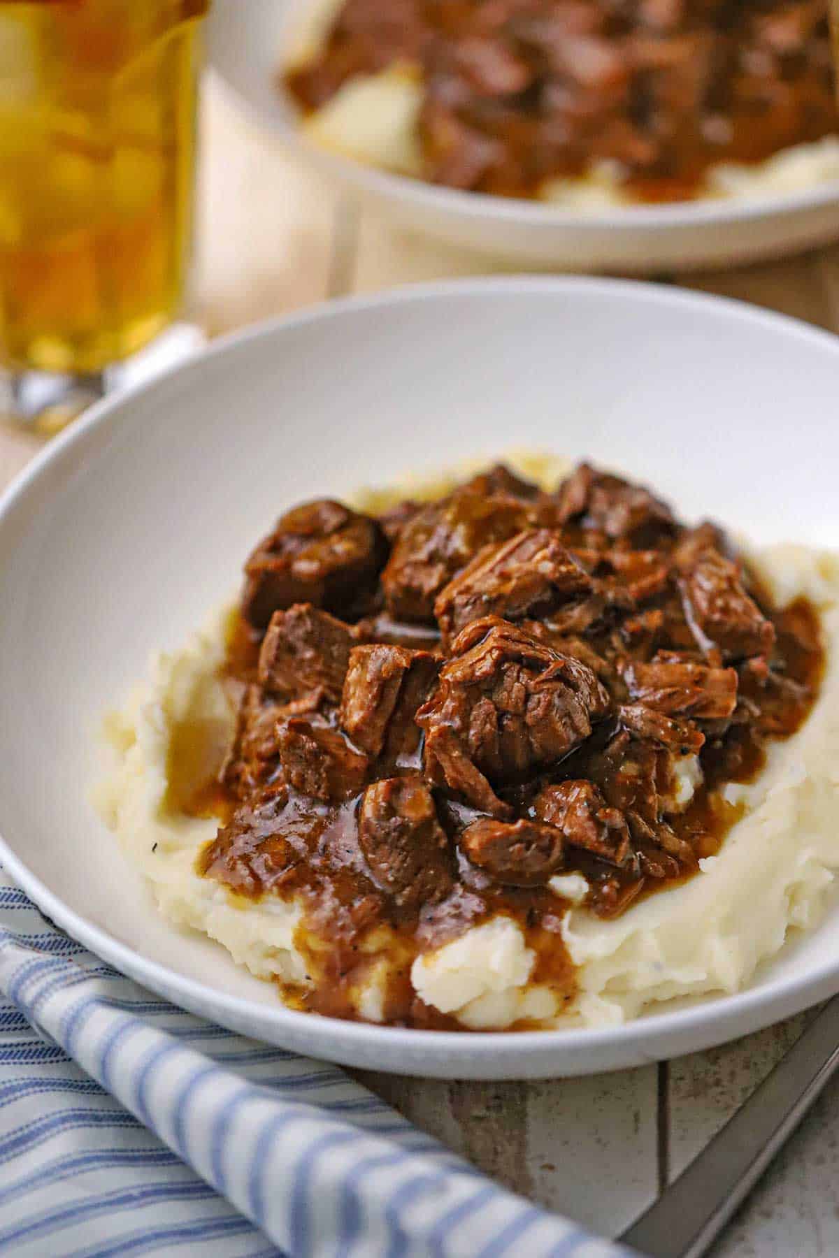 A shallow white bowl filled slow cooker beef tips and gravy over a pile of mashed potatoes with a glass of iced tea nearby.